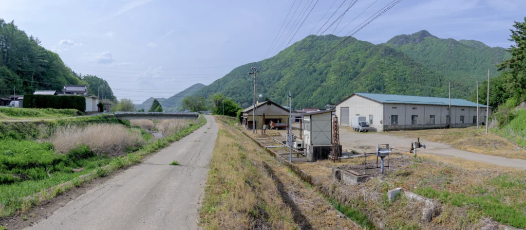 Panorama photograph of the village of Kawakami, showing a rural scenery with mountains in the background, and an agricultural building on the right side.